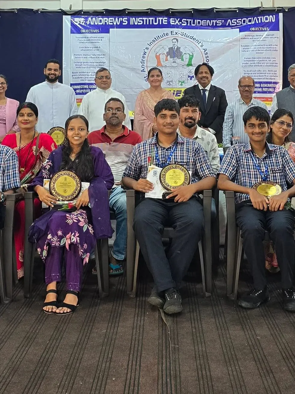 Students holding plaques at a SAIESA felicitation ceremony, with the St. Andrew's Institute Ex-Student's Association banner and committee members behind them