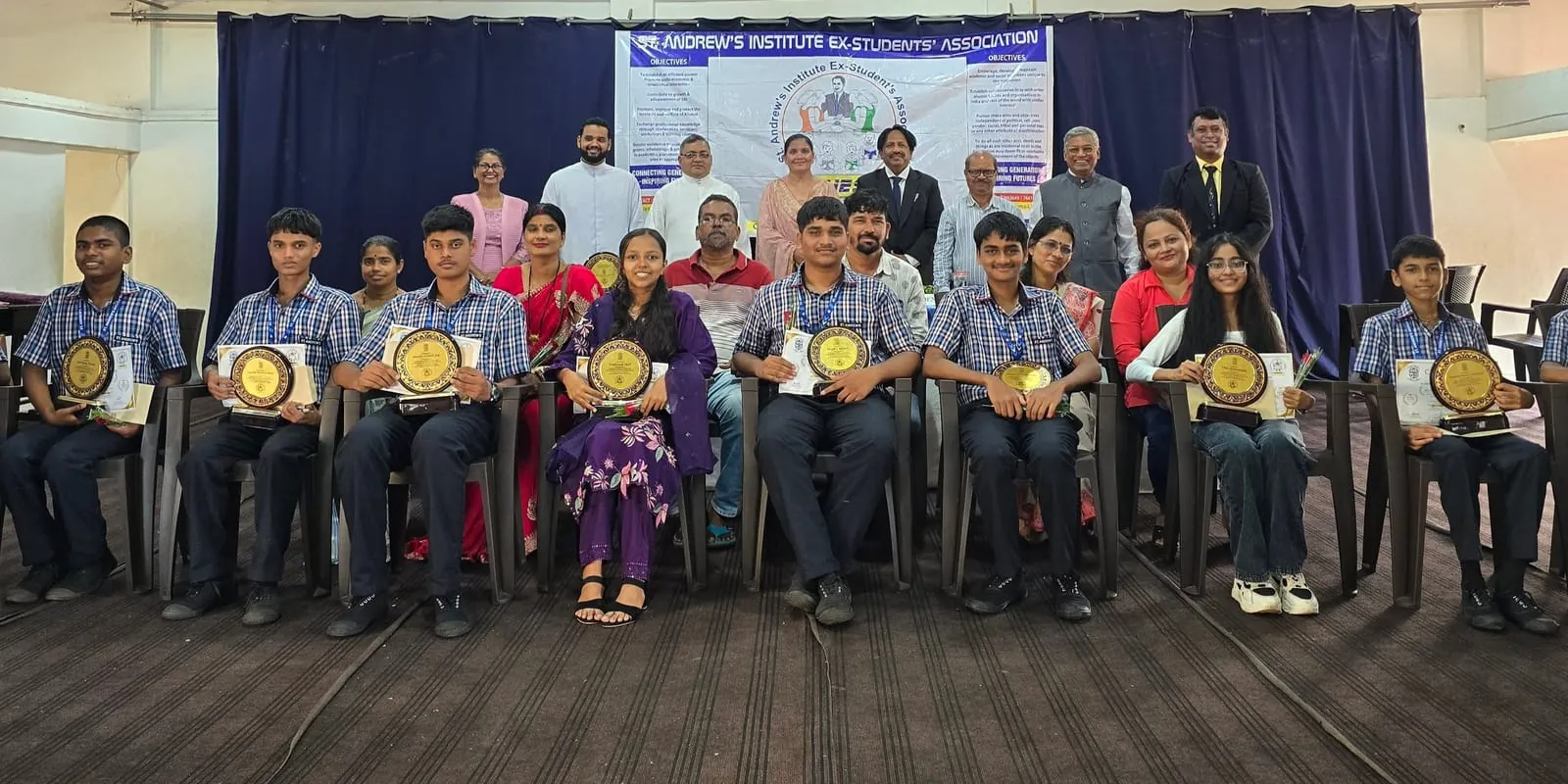 Students seated on stage holding plaques at a SAIESA felicitation ceremony, with committee members standing behind them and a St. Andrew's Institute Ex-Student's Association banner in the background