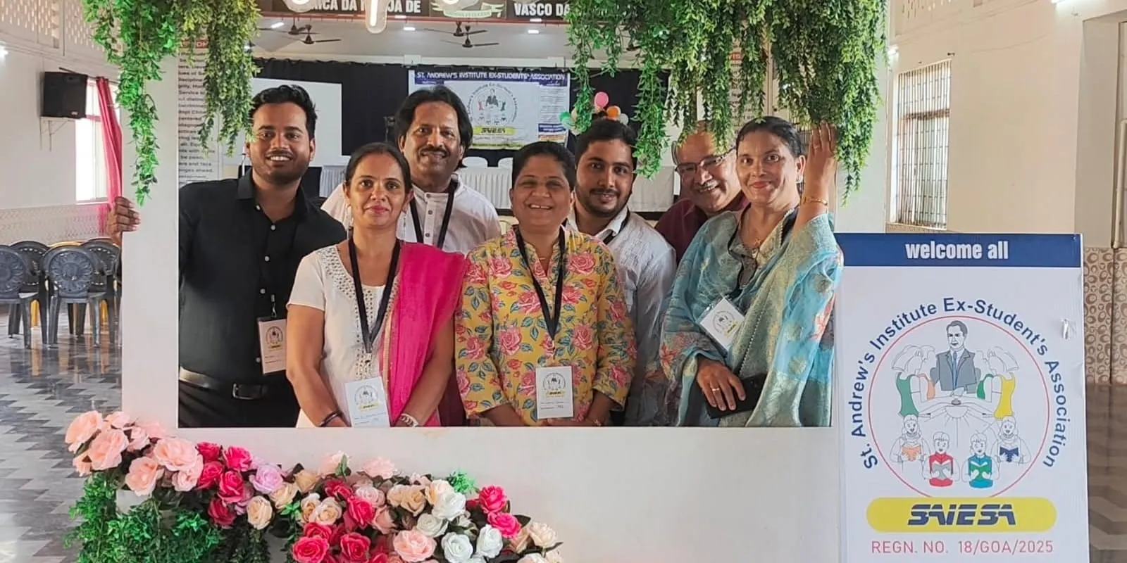 SAIESA committee and alumni members smiling together in a photo-booth frame decorated with flowers, next to a 'welcome all' St. Andrew's Institute Ex-Student's Association signboard